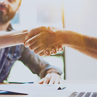 A photo graph of two people shaking hands with a man watching behind.